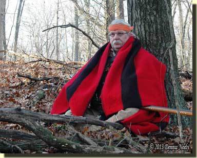 A traditional woodsman wrapped in a scarlet trade blanket sits against a red oak tree.