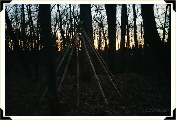 Bare log poles outline a peaked wigwam against a plain sunset.